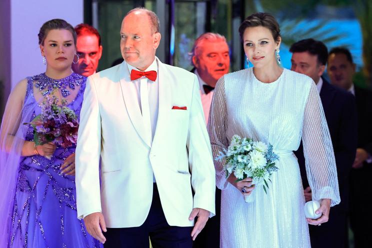 Prince Albert of Monaco in white tuxedo and red bow tie and Princess Charlene in a white dress holding flowers.